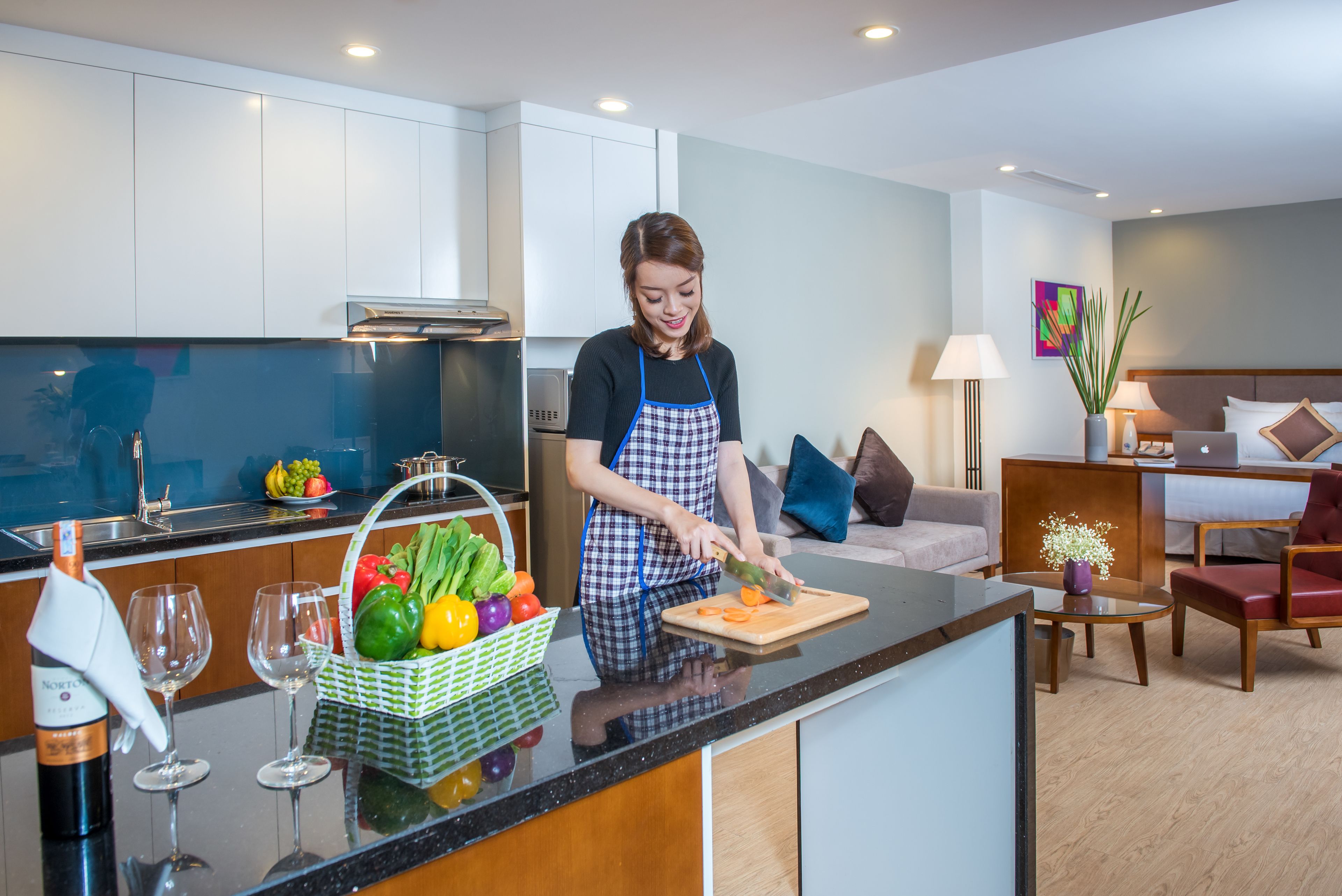 A modern kitchen with a person preparing food, a dining area with a sofa and table, and a living room with a bed and a lamp.