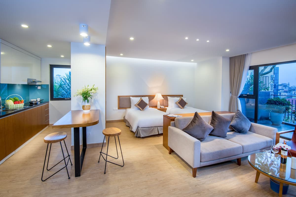 A modern hotel room with a kitchen area, a sofa, a dining table, and a large window with a city view.
