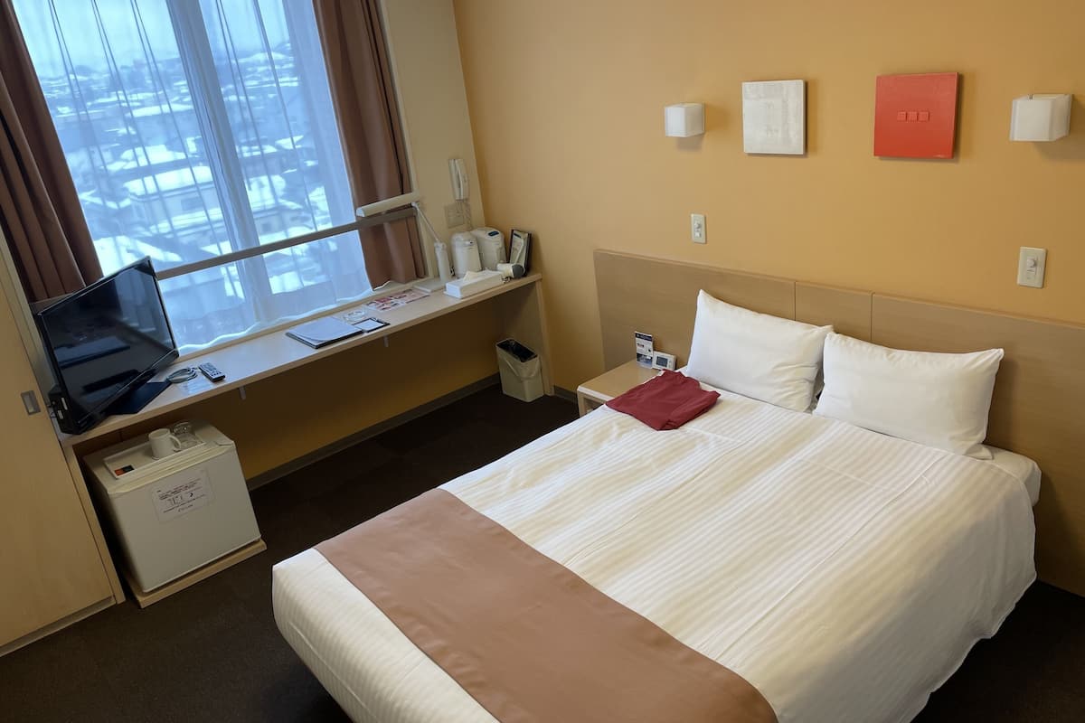 Hotellihuone, jossa on sänky, työpöytä tietokoneella, pieni roskakori ja ikkuna, jossa on verhot.