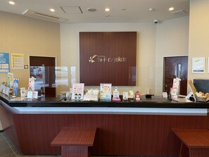 A hotel reception desk with a wooden counter, a large sign above it, and several informational brochures and a digital screen. - Quad Inn Yokote (Yokote)