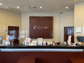 A hotel reception desk with a wooden counter, a large sign with the hotel name, and various informational brochures and amenities displayed. - Quad Inn Yokote (Yokote)