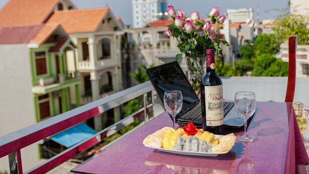 Una mesa con una computadora portátil, una botella de vino y un plato de fruta en un balcón con vistas a edificios.