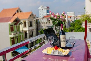 Una mesa con una computadora portátil, una botella de vino y un plato de fruta en un balcón con vistas a edificios.