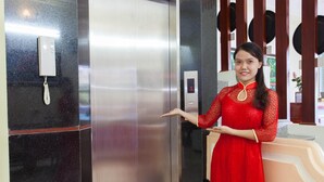 Una mujer con un vestido rojo está de pie junto a un ascensor, sosteniendo un teléfono y con una pantalla digital.