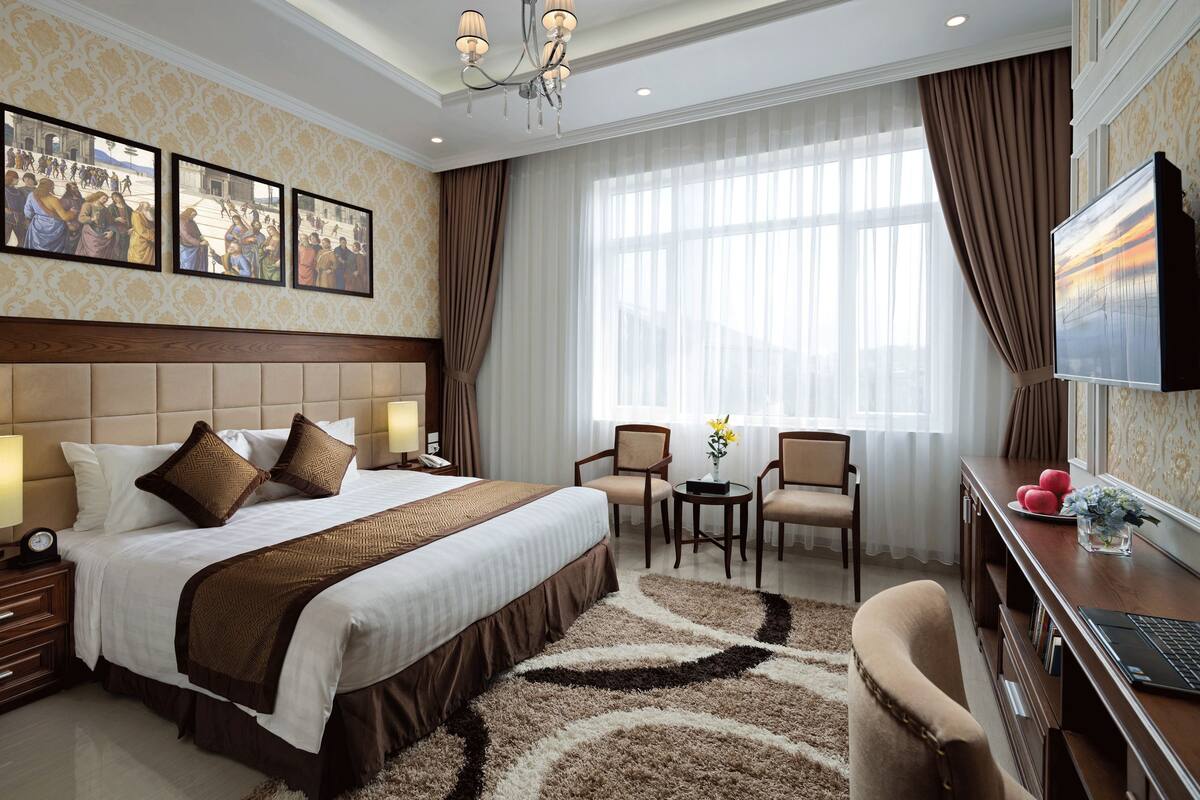 A hotel room with a large bed, two armchairs, a desk with a laptop, and a TV.