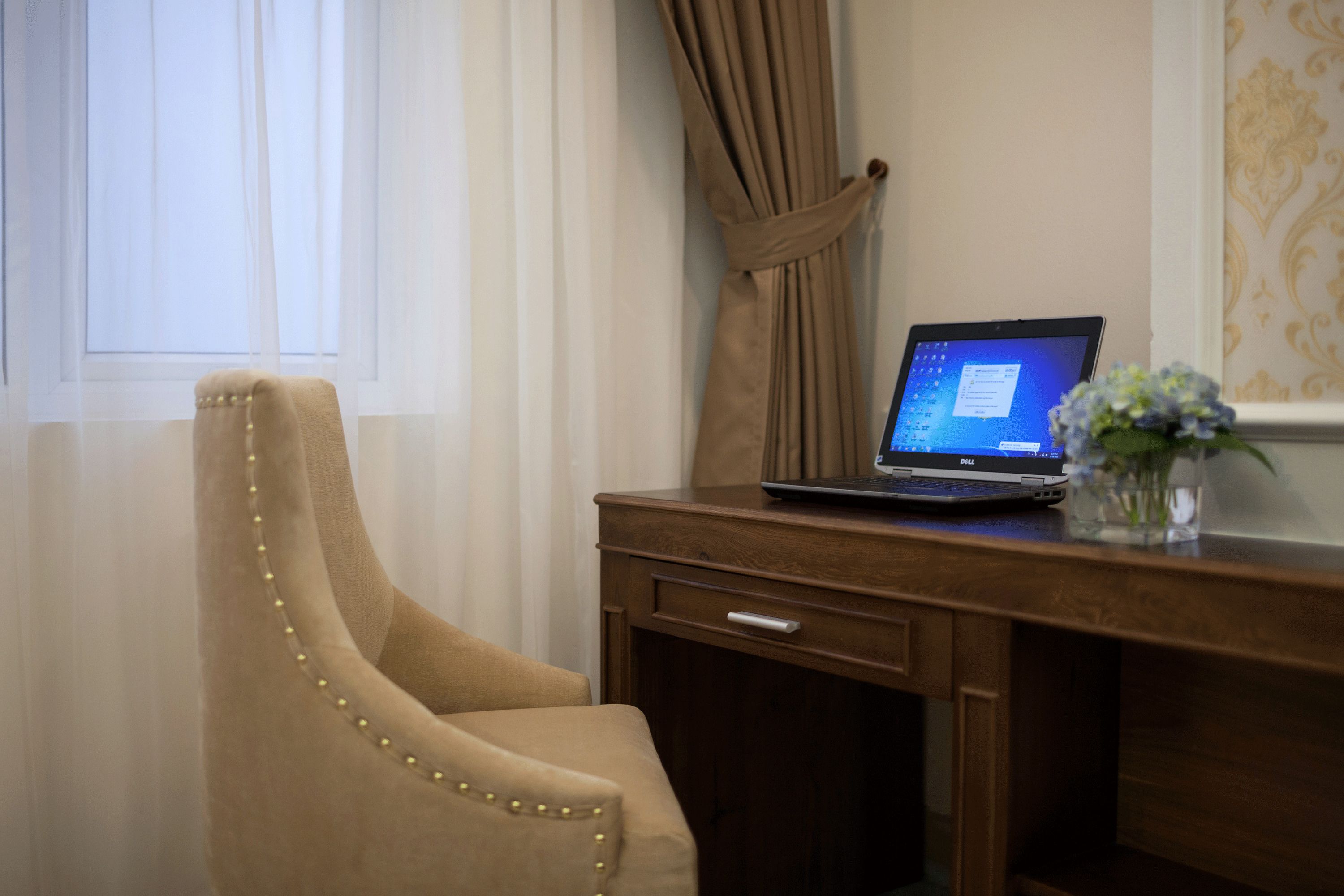 Un sillón beigüe con detalles perlados, un escritorio de madera con una computadora portátil y un florero con flores.