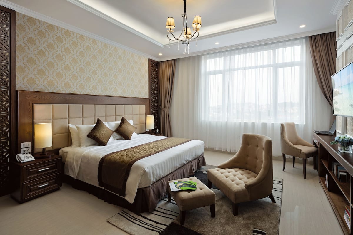 A spacious hotel room with a large bed, a sitting area with a chair and ottoman, a desk with a laptop, and a TV on the wall.