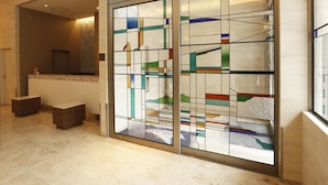 Um hall de hotel moderno com um balcão de recepção, um painel de vidro com padrões geométricos coloridos e piso de azulejos.
