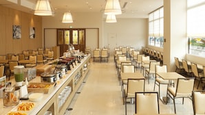 Ampla área de buffet com diversas mesas e cadeiras para refeições, balcão de serviço extenso com vários pratos e acompanhamentos, e janelas grandes que oferecem iluminação natural.