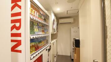 Un pasillo estrecho con una máquina expendedora que dice "KIRIN" a la izquierda, una unidad de aire acondicionado a la derecha y un refrigerador al fondo.