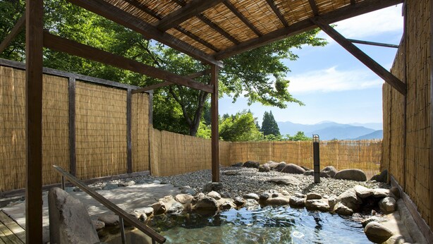 A traditional Japanese onsen with a wooden structure, a stone bath, and a view of mountains in the background. - Muica Onsen Hotel (Minamiuonuma)