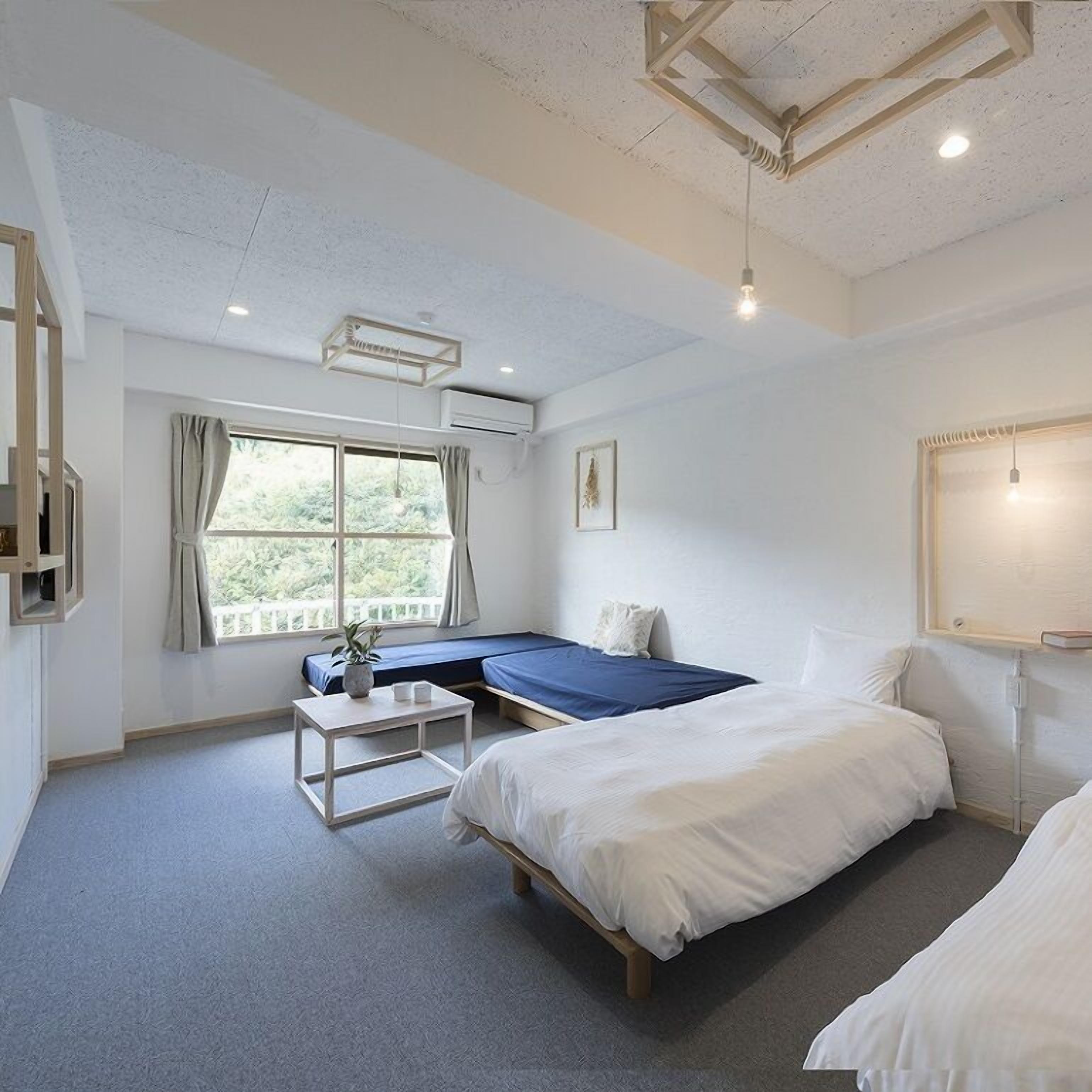 Ett sovrum med två sängar, ett litet bord, ett fönster med gardiner och en taklampa.
