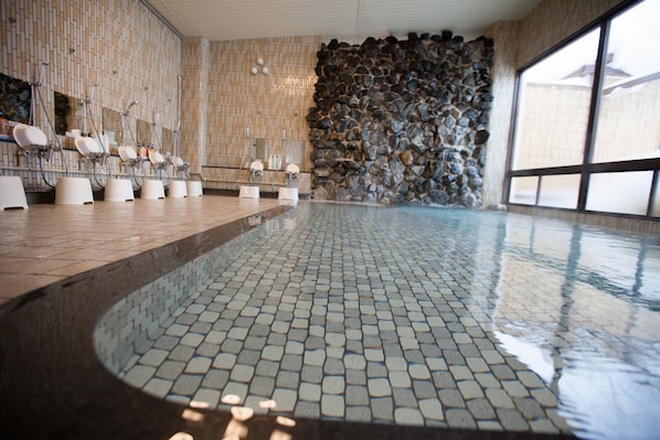 A modern indoor swimming pool with tiled flooring, multiple changing stations, and a stone wall. - Muica Onsen Hotel (Minamiuonuma)