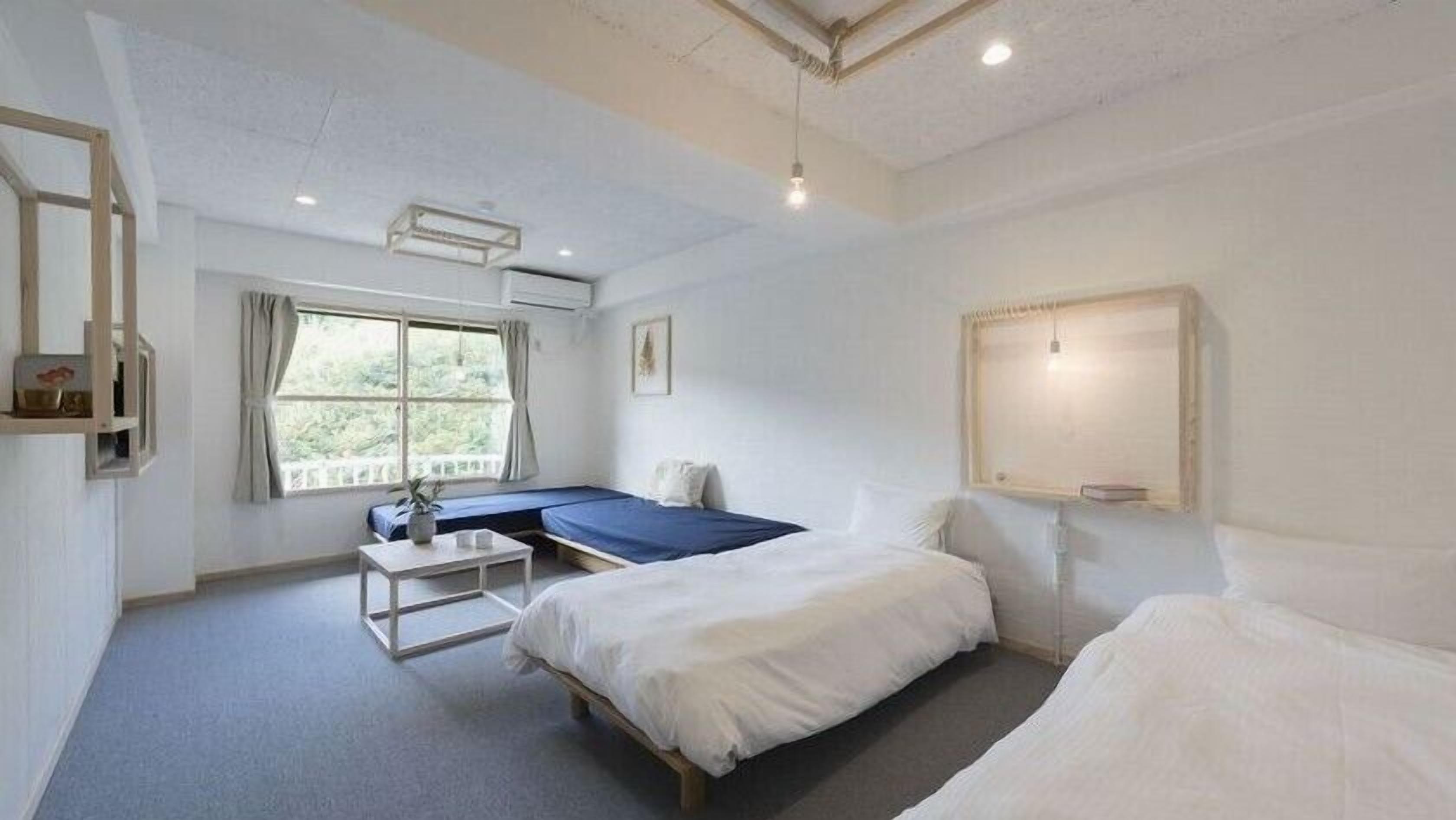 A room with two beds, a small table, a window with curtains, and a wall-mounted light fixture.