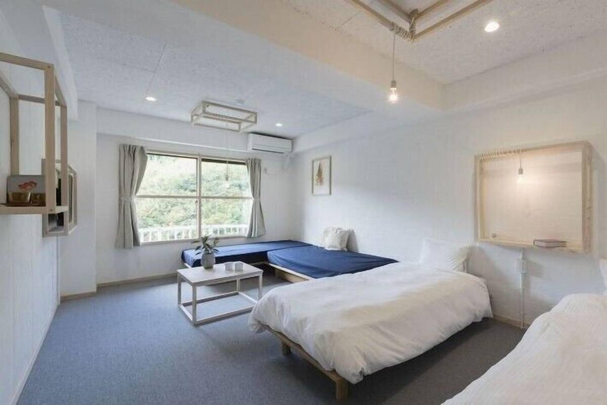 A room with two beds, a small table, a window with curtains, and a wall-mounted light fixture.