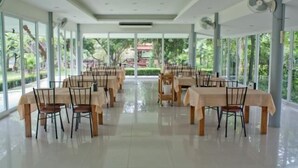 A spacious indoor dining area with multiple tables and chairs, large windows offering a view of greenery outside. - Ayodhara Village (Ayutthaya)