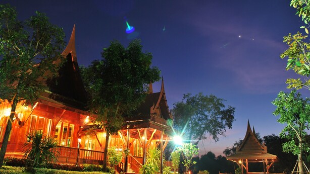 A traditional wooden building with a thatched roof, surrounded by lush greenery and illuminated by warm lighting. - Ayodhara Village (Ayutthaya)