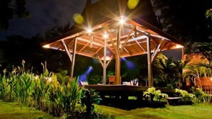 A wooden pavilion with a roof supported by beams, surrounded by greenery and plants. - Ayodhara Village (Ayutthaya)