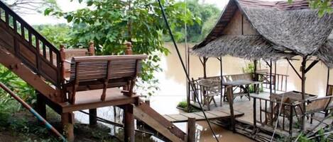 A wooden deck with a staircase leading to a raised platform, a thatched-roof structure, and a dining area with chairs and tables.