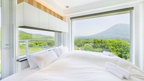 Habitación de hotel moderna con una cama grande, ropa de cama blanca y vistas a una montaña y vegetación.