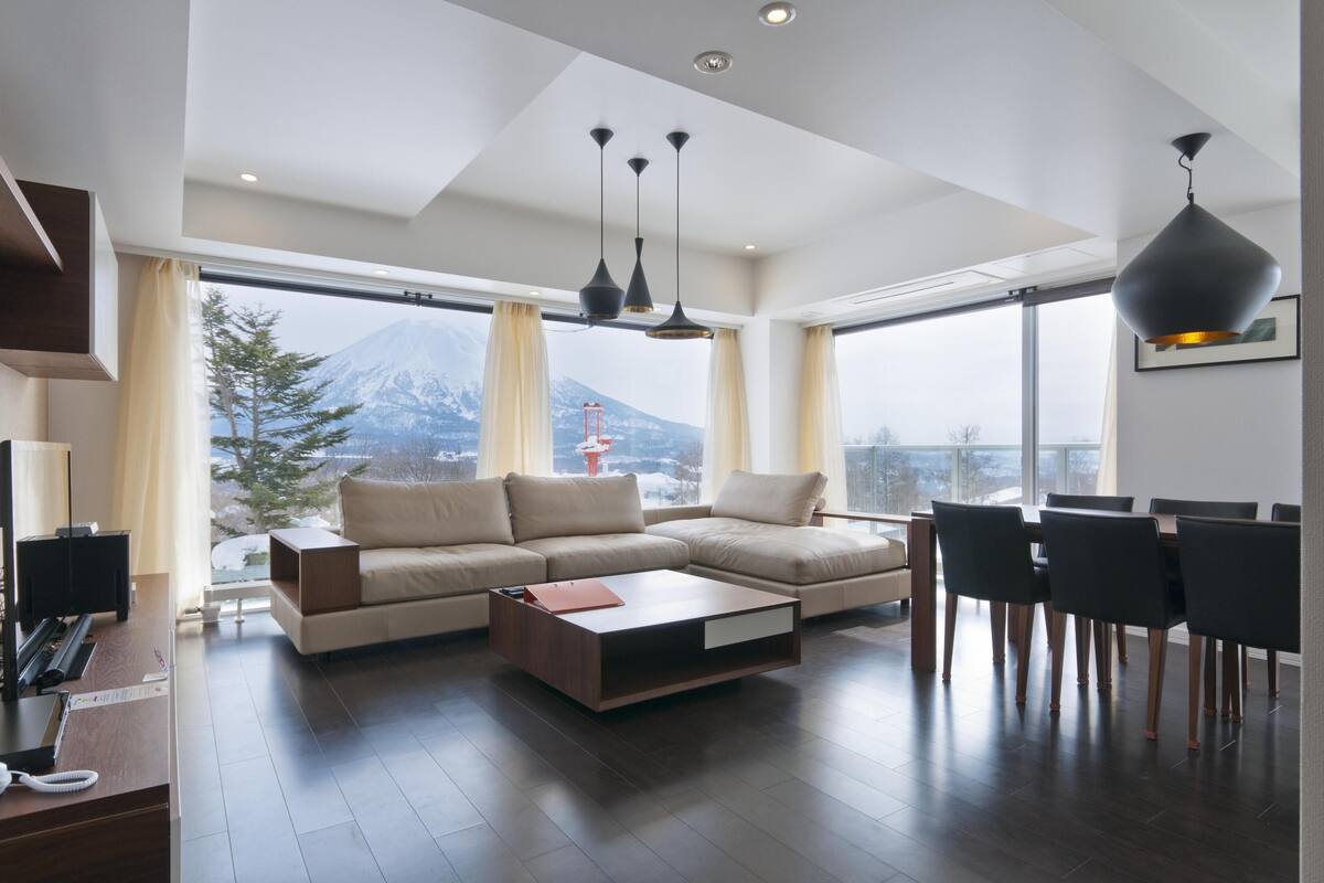 A modern living room with a large sofa, a coffee table, and a dining area with chairs. There are large windows with a view of a mountain and a crane.