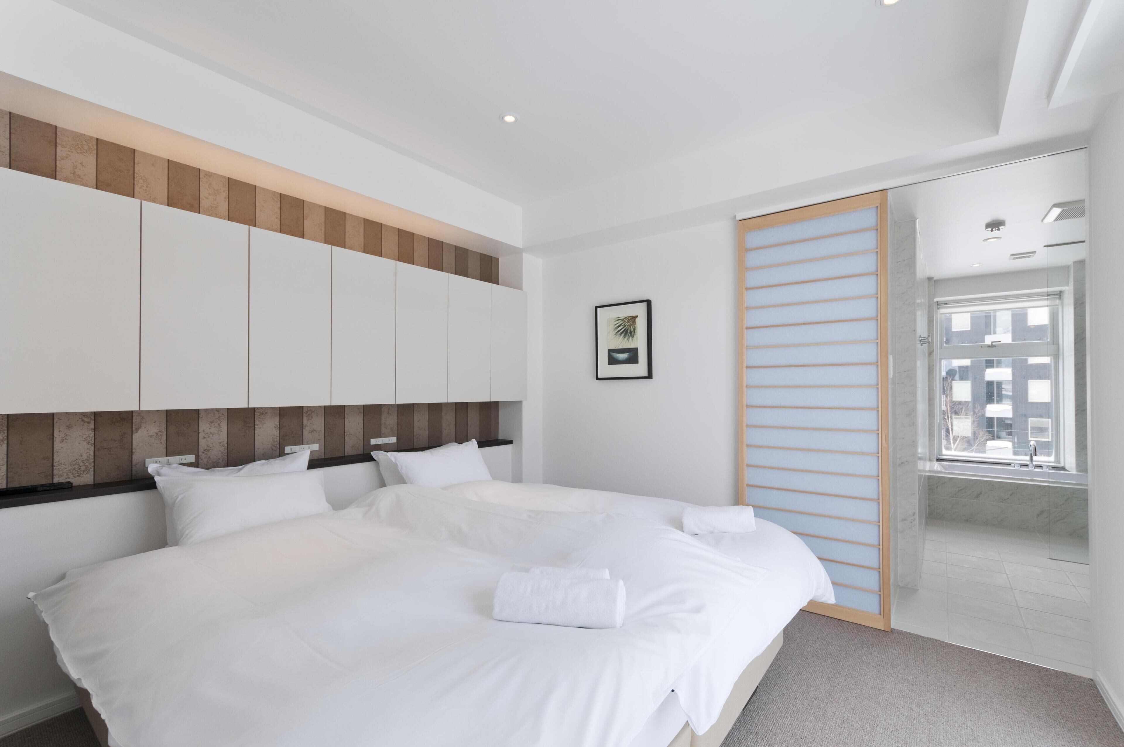 A modern hotel room with a large bed, a wooden headboard, a glass shower enclosure, and a view of the city through the window.