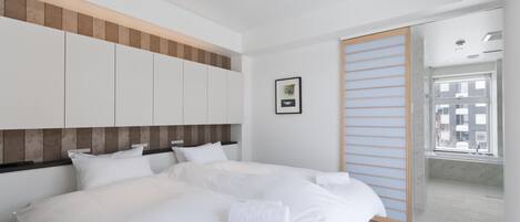 A modern hotel room with a large bed, a wooden headboard, a glass shower enclosure, and a view of the city through the window.