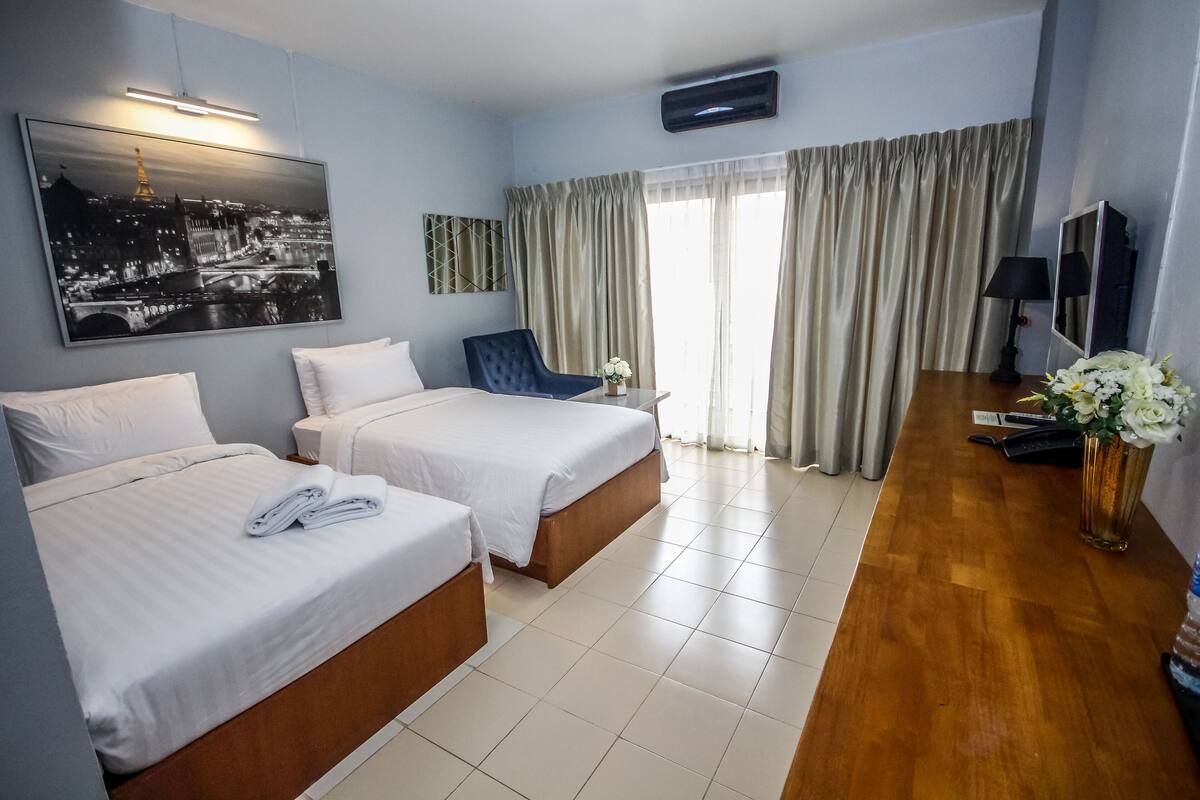A hotel room with two beds, a desk, a chair, a TV, and a large window with curtains.