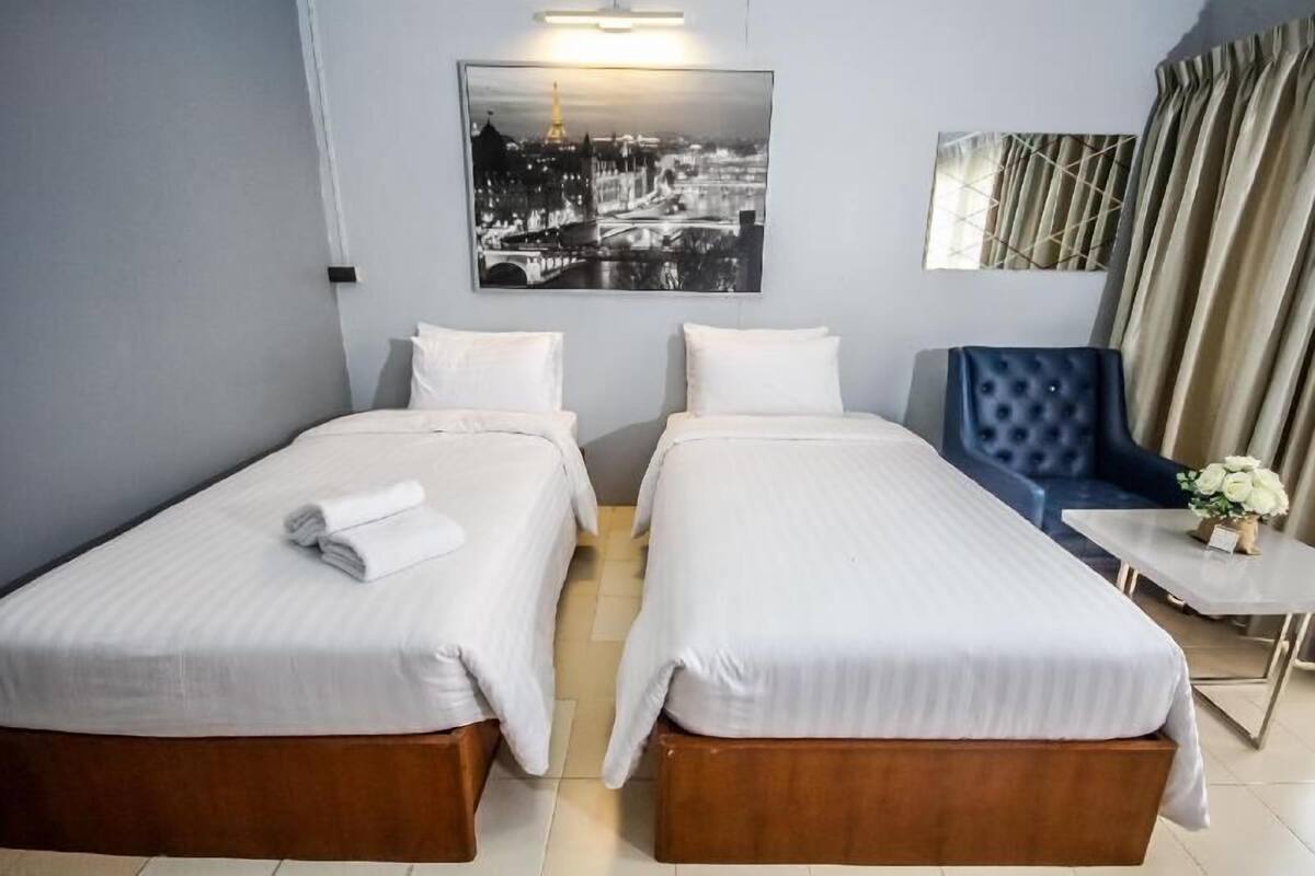 A hotel room with two beds, a blue armchair, a small table with a vase, and a cityscape painting on the wall.