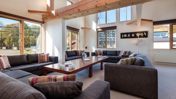 A spacious living area with multiple sofas and a central coffee table, large windows providing natural light, and a wooden ceiling structure. - Gondola Chalets (Kutchan)