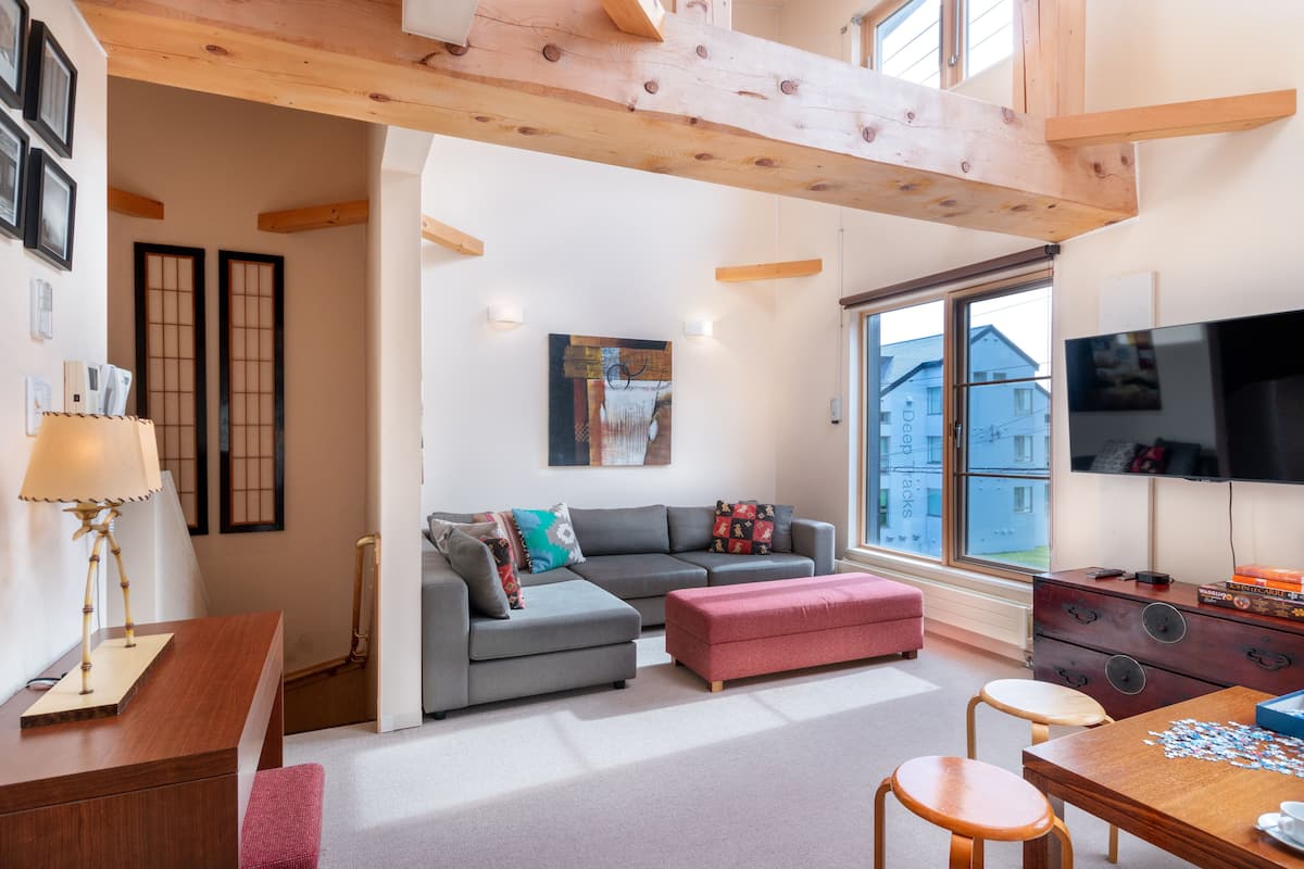 Un salon moderne avec un canapé gris, un pouf rouge, une table basse en bois et un téléviseur posé sur un meuble en bois.