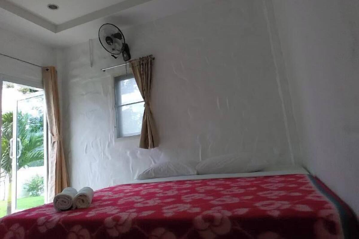 Un dormitorio con cama, ventana con cortinas, ventilador de techo y vistas a zonas verdes.