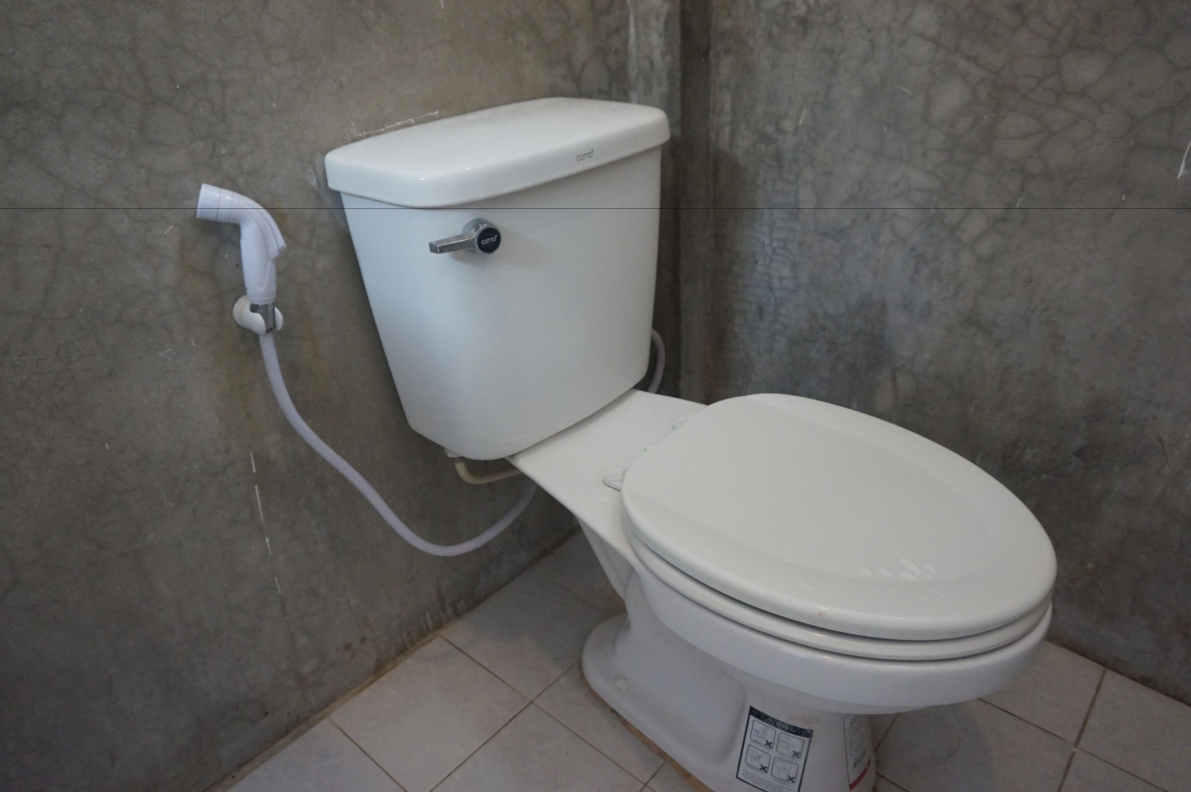 A white toilet with a control knob and a flush handle, connected to a wall-mounted pipe.