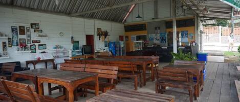 Un espace extérieur abrité avec des tables et des bancs en bois, un tableau noir servant de menu et une poubelle bleue.