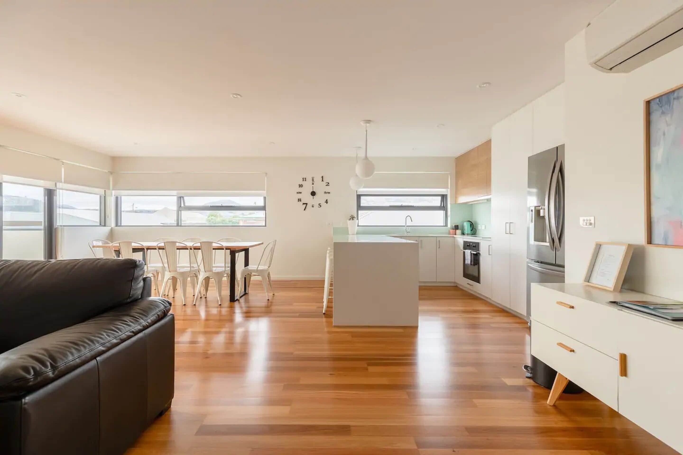 A modern living room with a black sofa, a dining area with white chairs, a kitchen with white cabinets, and a large window providing natural light.