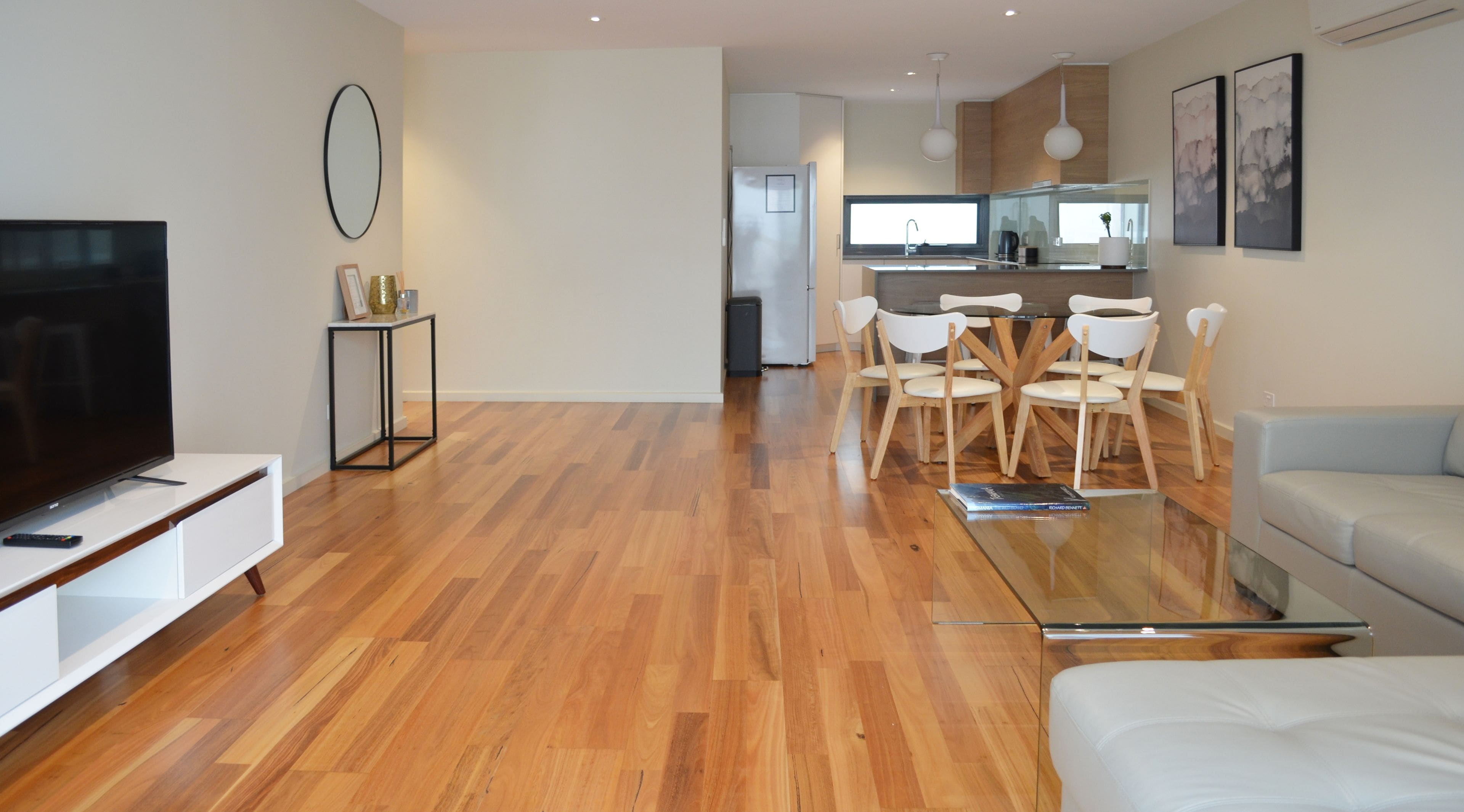 A modern living room with a flat-screen TV, a glass coffee table, and a sofa. There is a dining area with a wooden table and chairs. The room has wooden flooring and white walls.