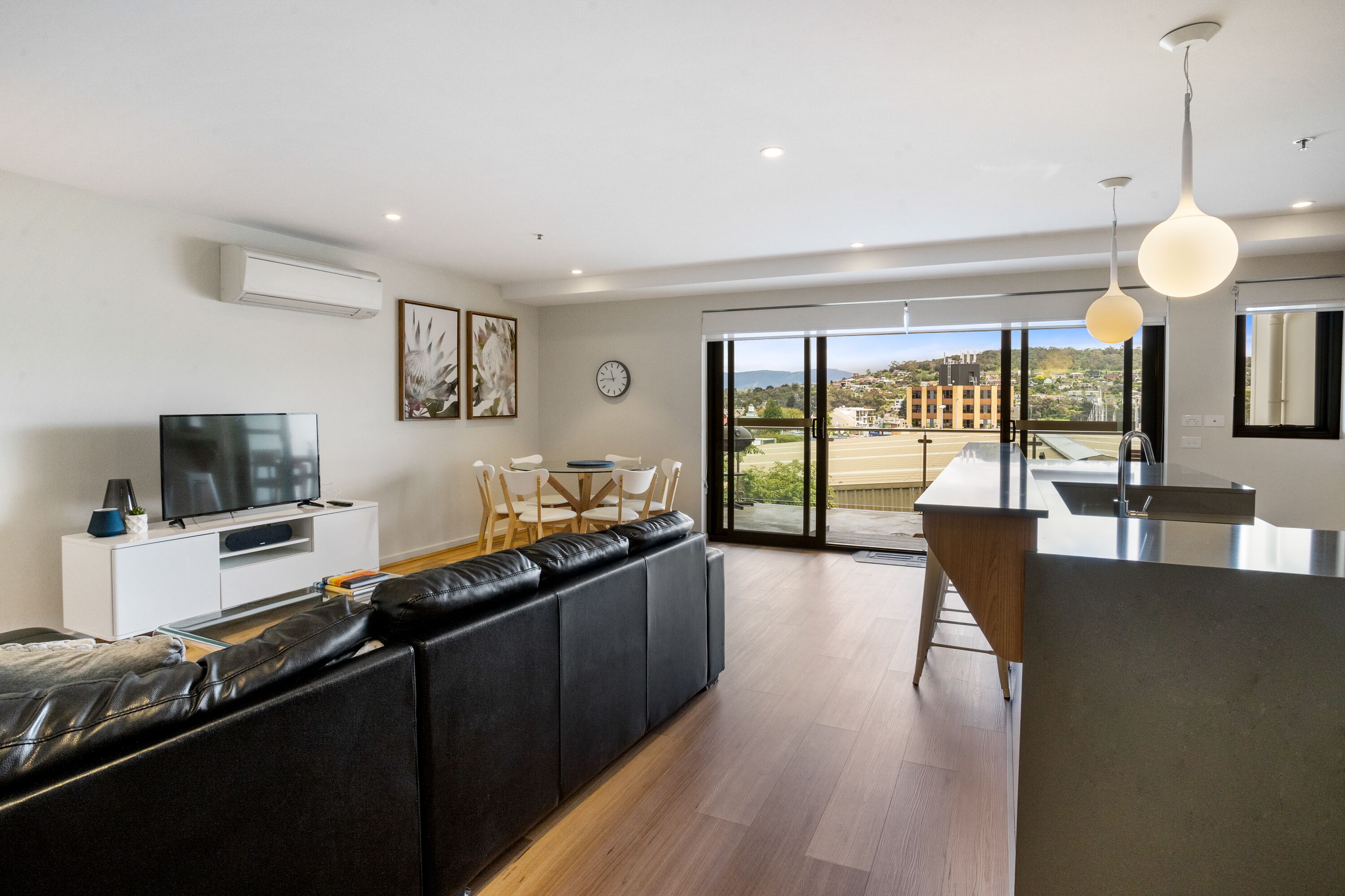 A modern living room with a black leather sofa, a flat-screen TV, a dining area with a table and chairs, and a kitchen area with a large island.
