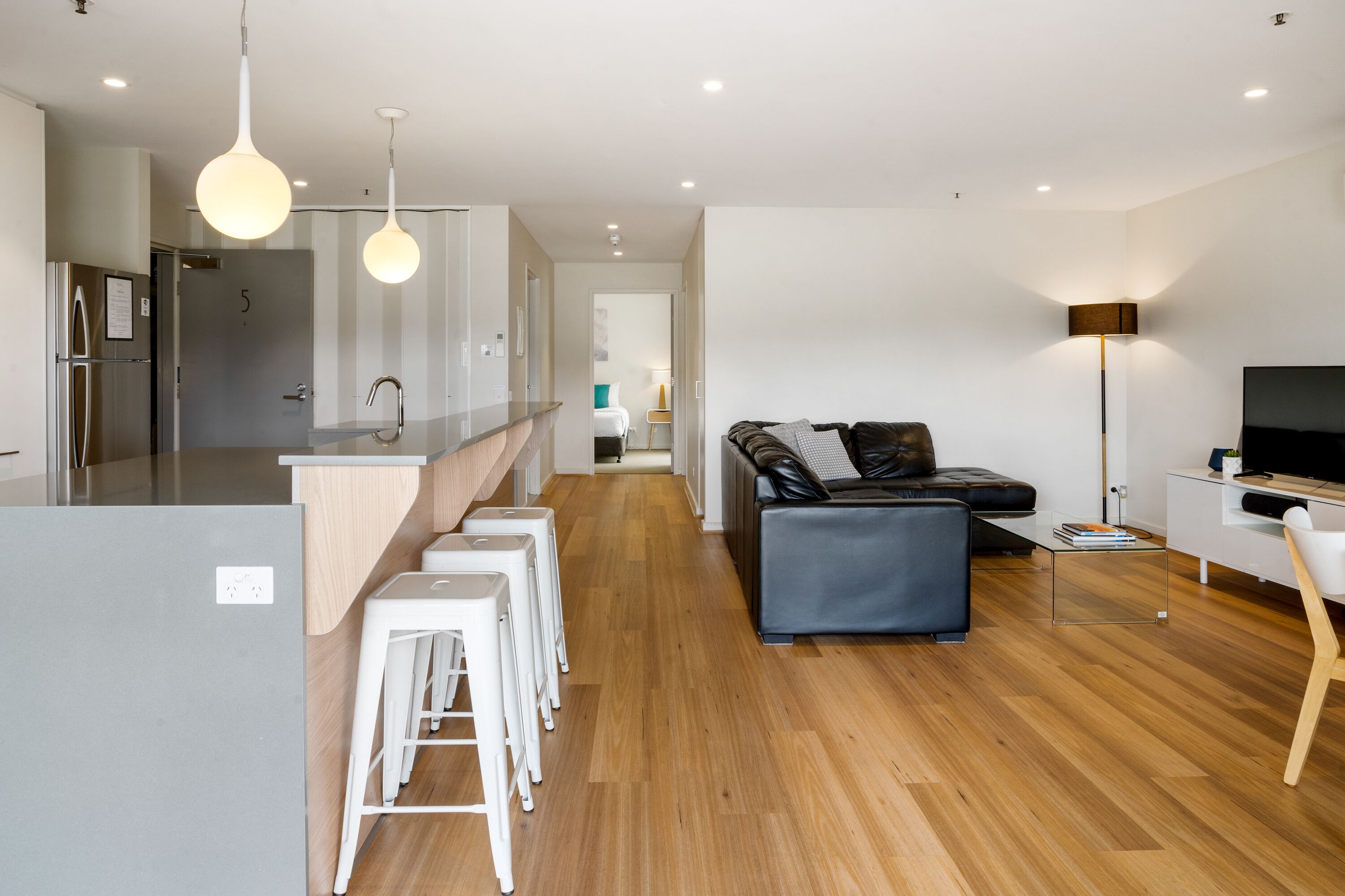 A modern kitchen with white bar stools, a sofa, and a TV in a living area with wooden flooring.