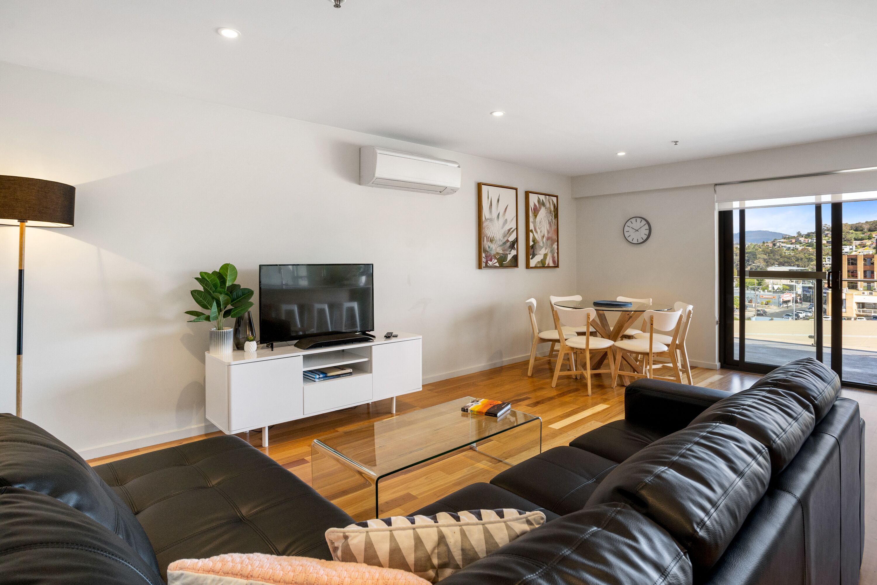 Een moderne woonkamer met een zwarte leren bank, een glazen salontafel, een televisie op een witte kast, en een eethoek met een tafel en stoelen.