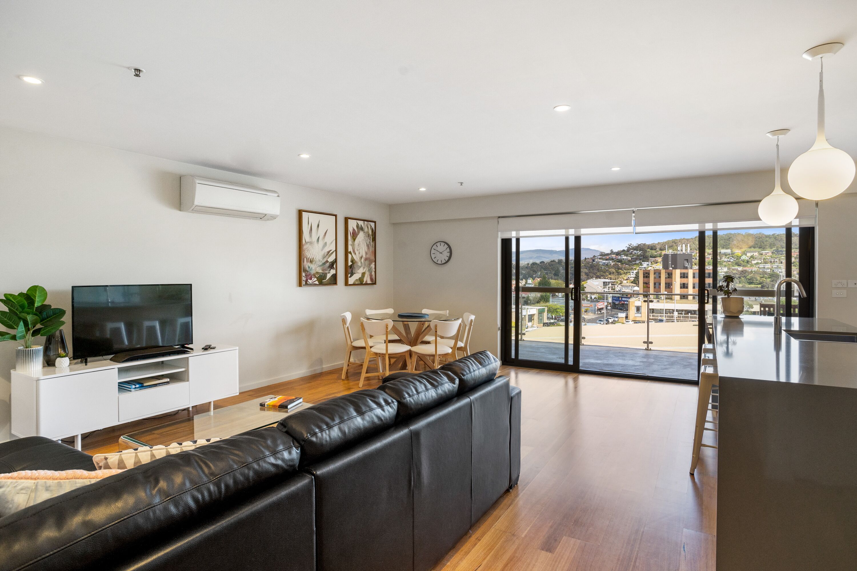 Un salon moderne avec un canapé en cuir noir, un téléviseur à écran plat, un coin repas comprenant une table et des chaises, et une grande porte-fenêtre coulissante donnant sur un balcon avec vue sur la ville.
