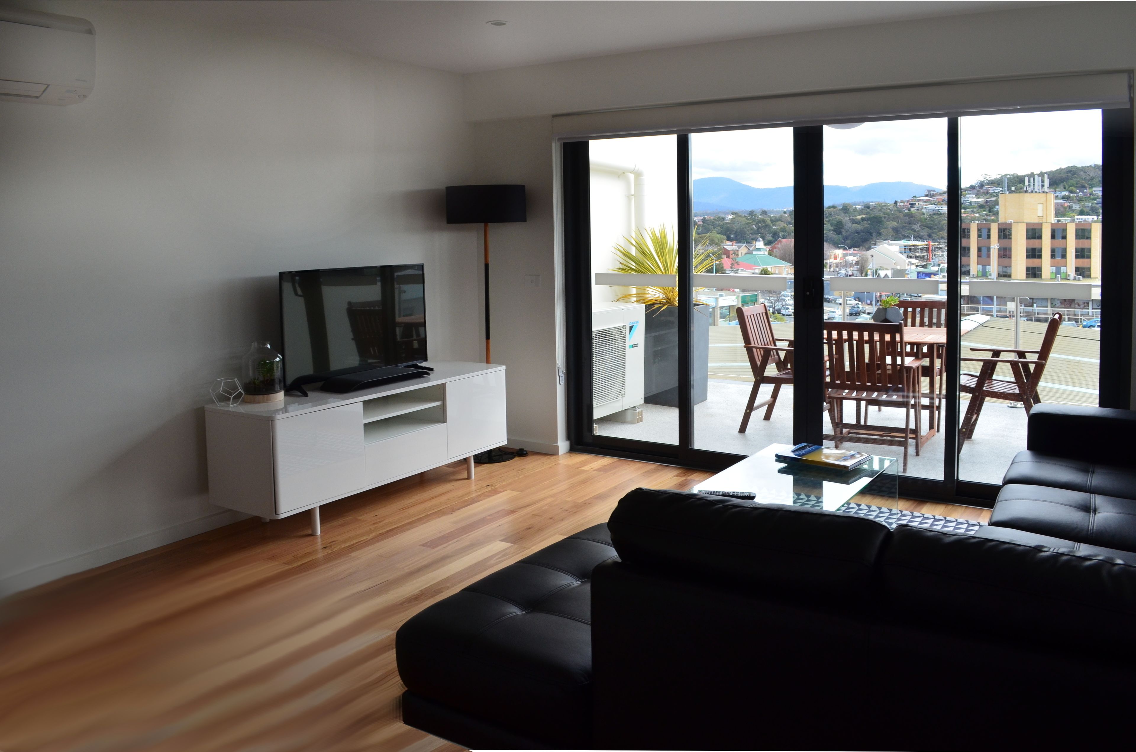 A modern living room with a black sofa, a flat-screen TV on a white console, a dining area with wooden chairs, and a large sliding glass door leading to an outdoor balcony with a view of the city and mountains.
