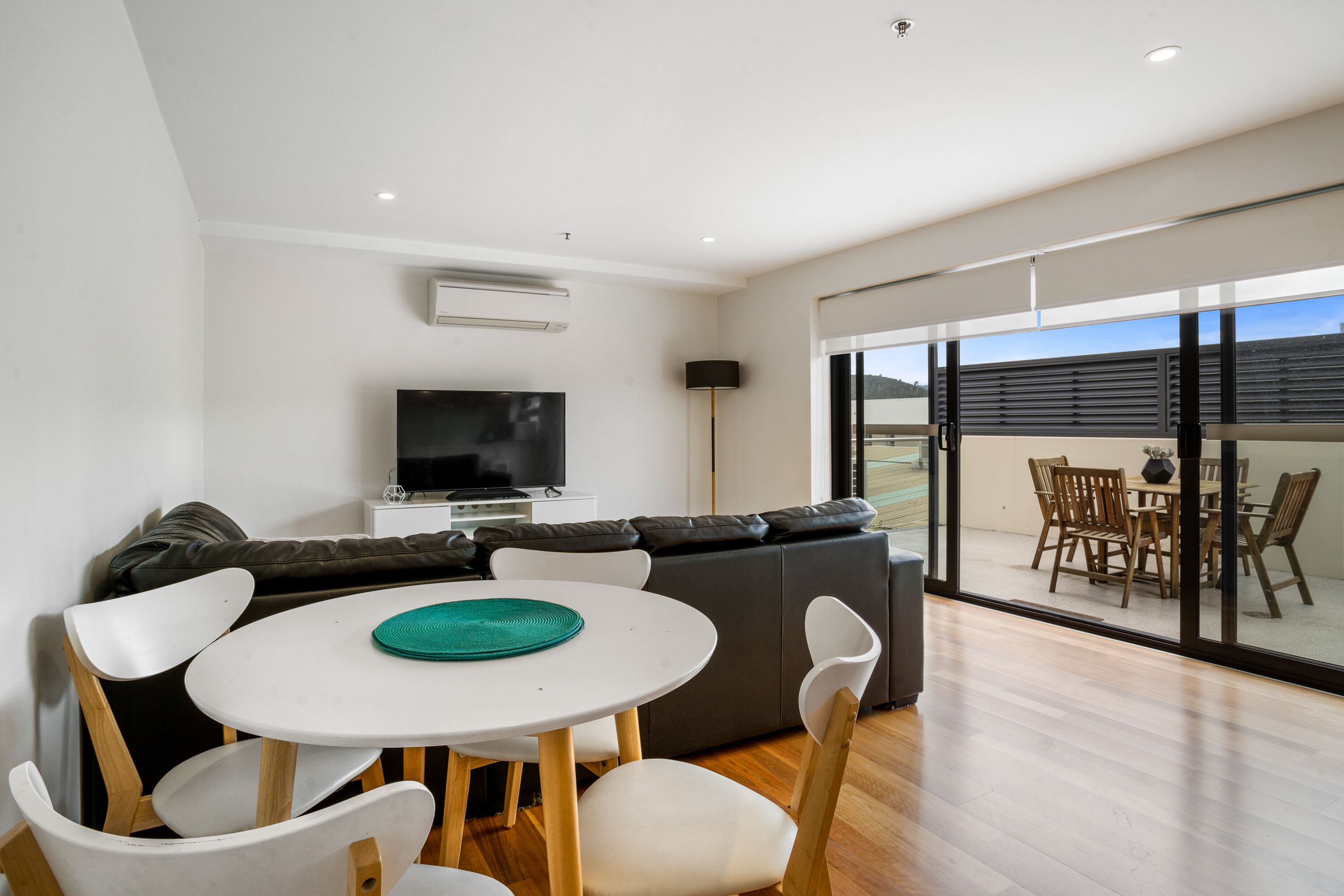 A modern living room with a white round table, white chairs, a grey sofa, a flat-screen TV, and a balcony with wooden furniture.