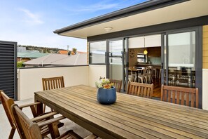A wooden table with chairs and a blue vase, a sliding glass door leading to a kitchen, and a view of neighboring houses. - Kangaroo Bay Apartments (Bellerive)