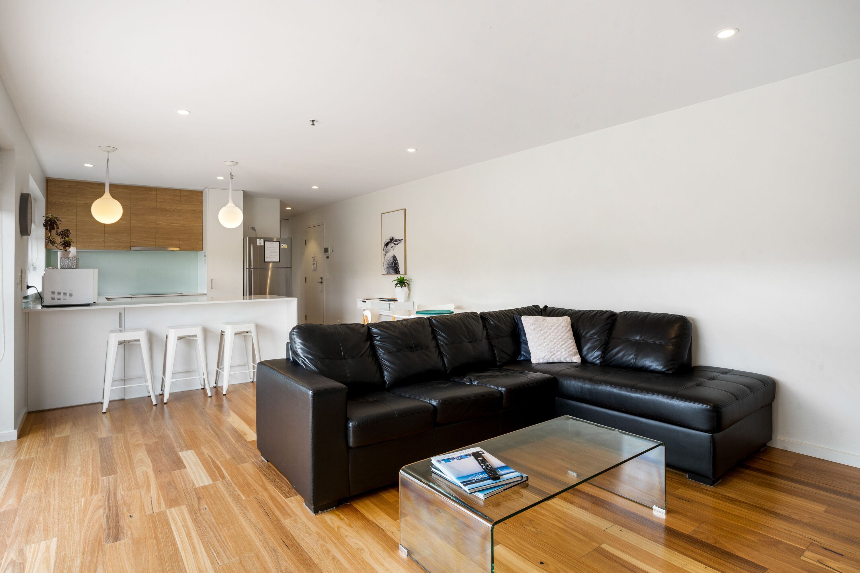 A modern living room with a black leather sectional sofa, a glass coffee table, and a kitchen area with white bar stools.