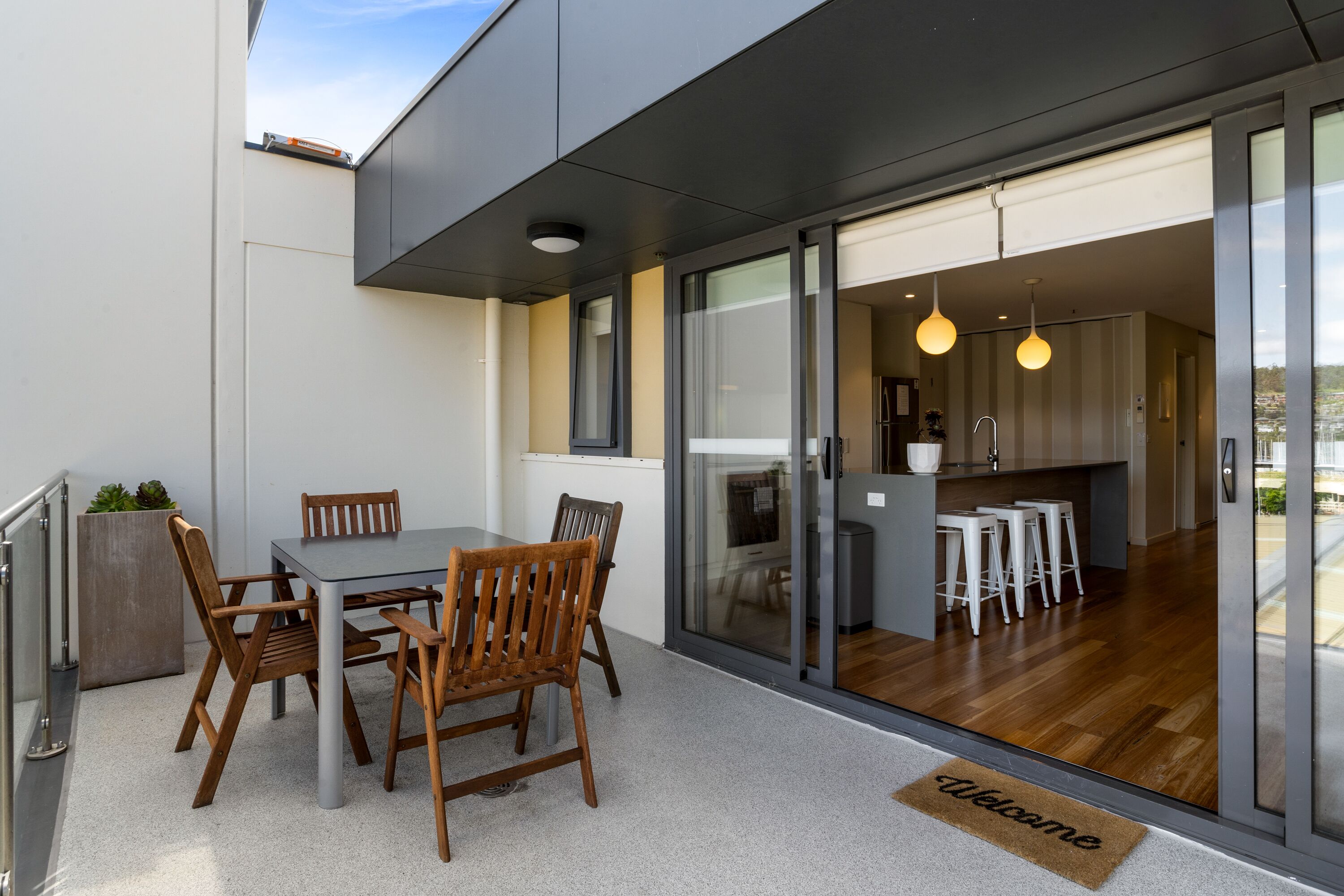 A modern outdoor dining area with a table and chairs, a welcome mat, and a view into the kitchen and living space.