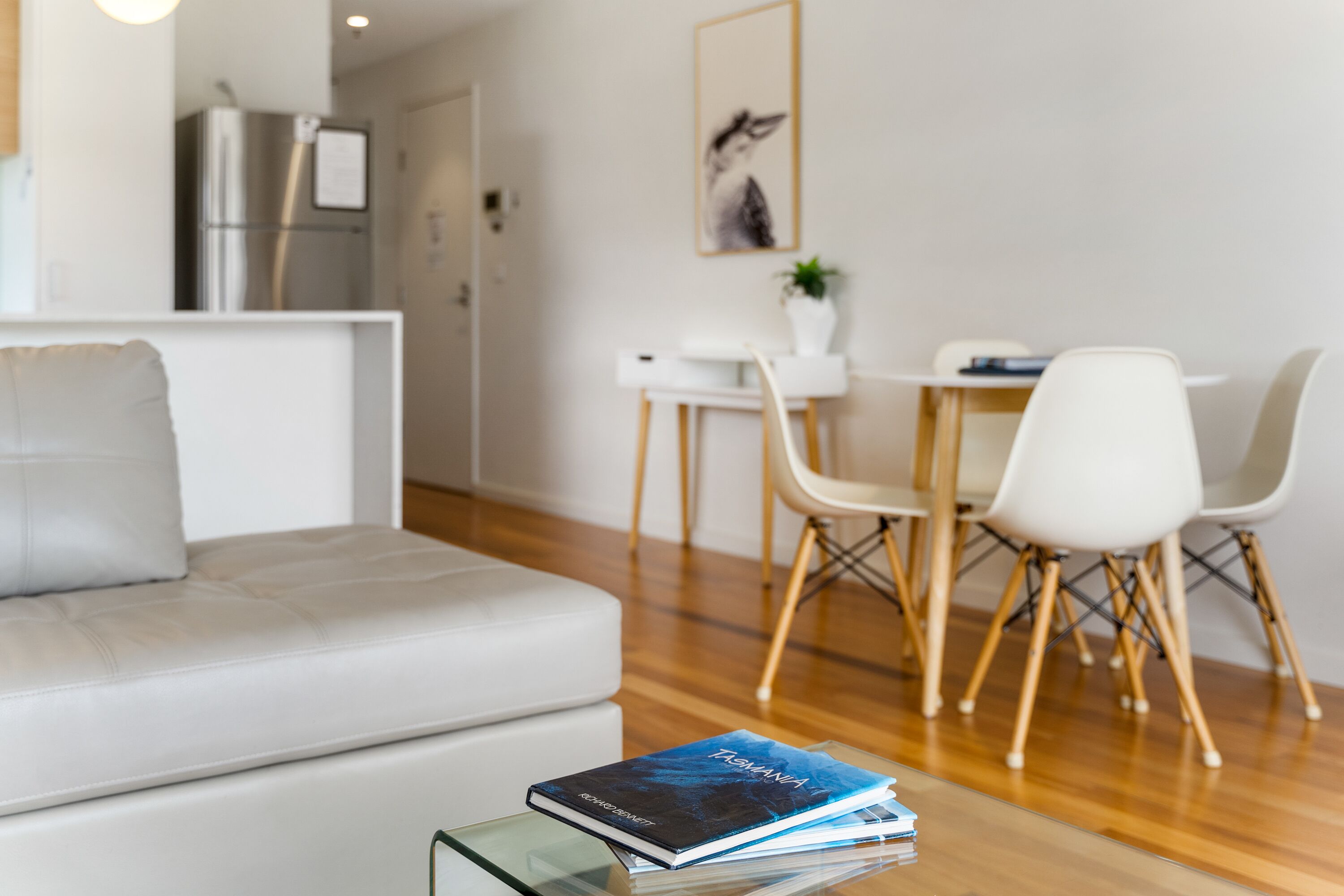 Uma sala de estar moderna com sofá, uma mesa de centro de vidro com livros, um espaço de jantar com mesa e cadeiras brancas, e uma cozinha com geladeira.