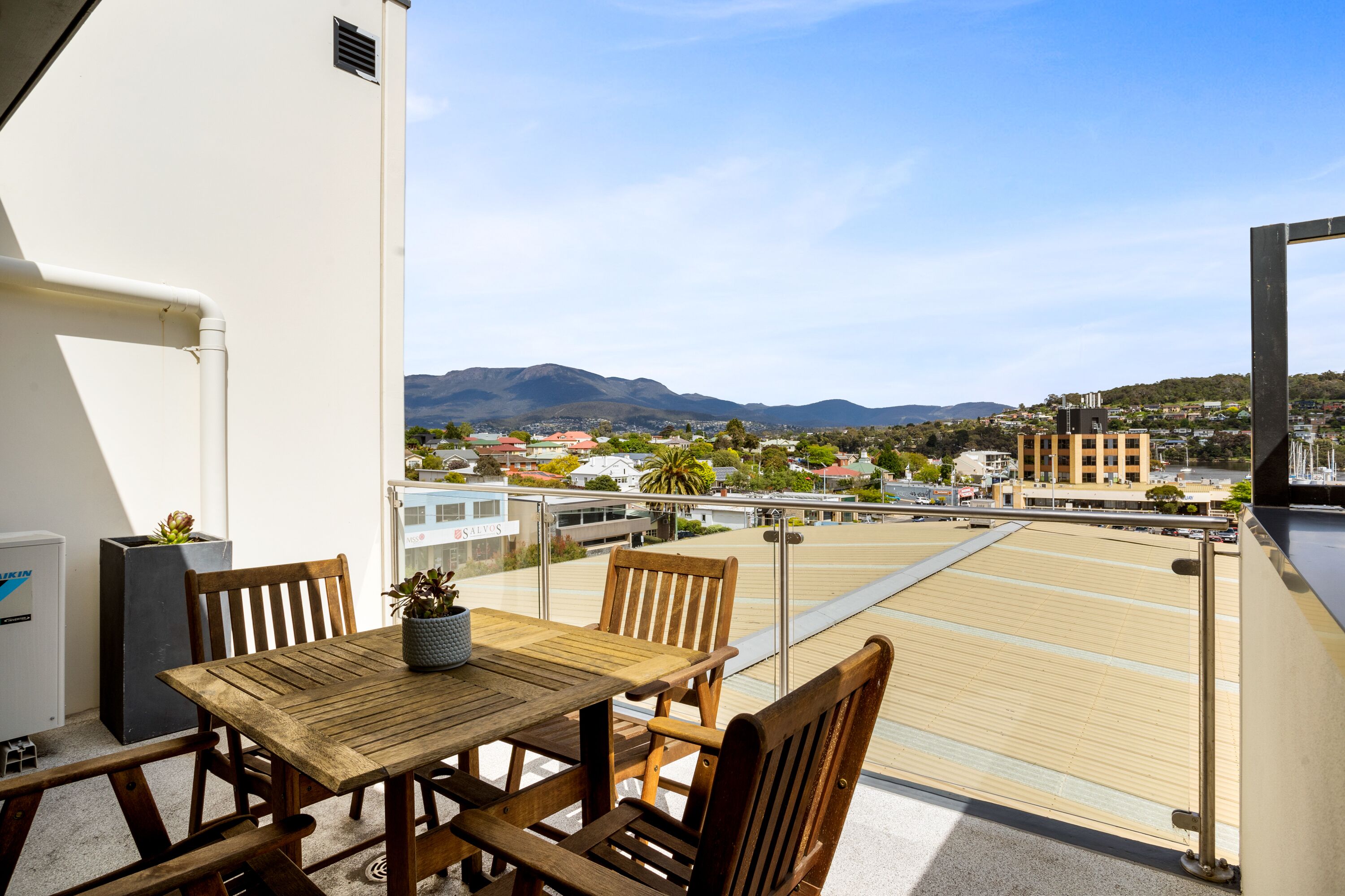 Uma varanda com mesa e cadeiras de madeira, um vaso com planta e vista para uma paisagem urbana com montanhas ao fundo.
