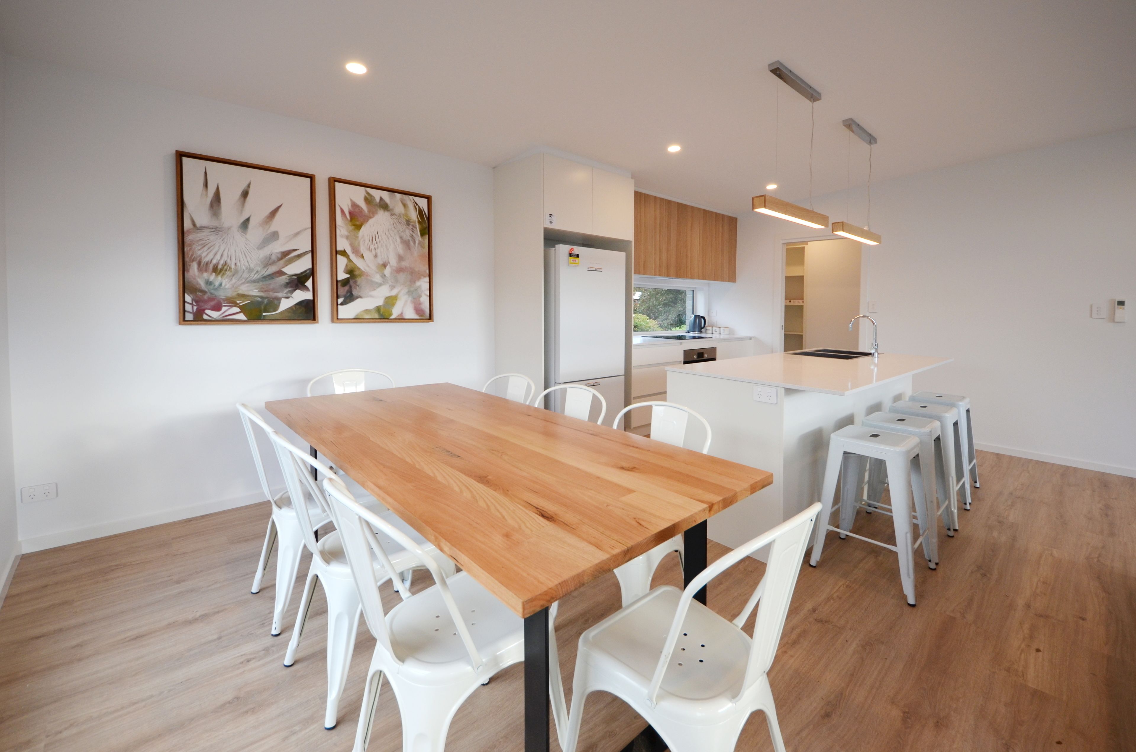 A modern kitchen with white cabinets, a wooden dining table, and white chairs. There are two framed floral paintings on the wall.