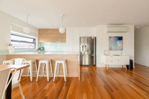 A modern kitchen with wooden flooring, white bar stools, a stainless steel refrigerator, and a minimalist white cabinet. - Kangaroo Bay Apartments (Bellerive)