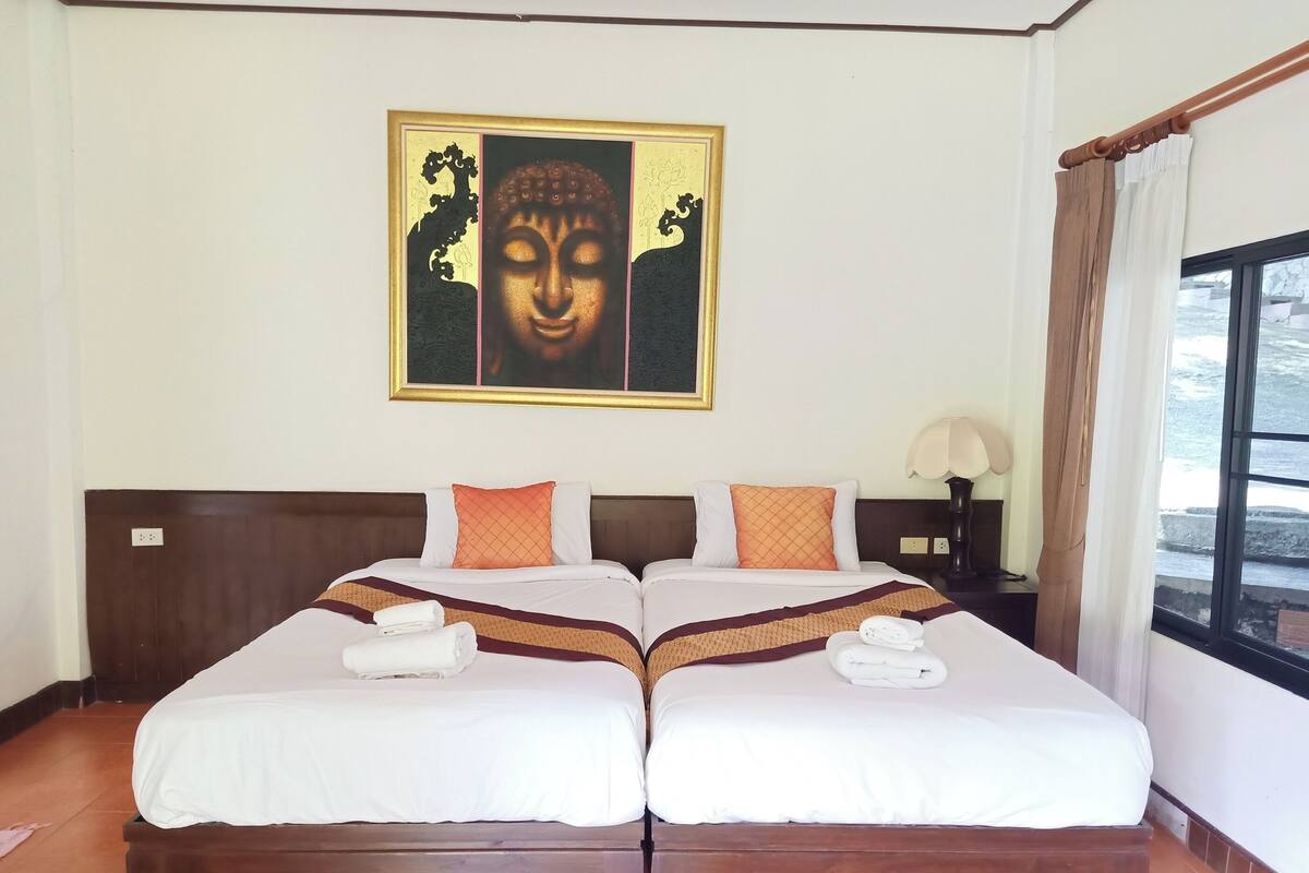 Una cama doble con sábanas blancas y dos almohadas naranjas, una mesita de noche con una lámpara, y un cuadro de Buda en la pared.
